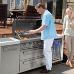 A modern Mont Alpi modular outdoor kitchen setup. From left to right, the modules include an infrared side burner, a large 304 stainless steel gas grill with an open hood showcasing internal halogen lights, an outdoor sink, and a glass-door beverage refrigerator. The entire island features bright white granite countertops and sits on a contemporary backyard wooden deck at dusk.