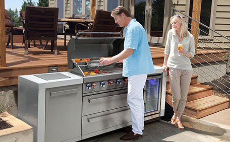 A modern Mont Alpi modular outdoor kitchen setup. From left to right, the modules include an infrared side burner, a large 304 stainless steel gas grill with an open hood showcasing internal halogen lights, an outdoor sink, and a glass-door beverage refrigerator. The entire island features bright white granite countertops and sits on a contemporary backyard wooden deck at dusk.